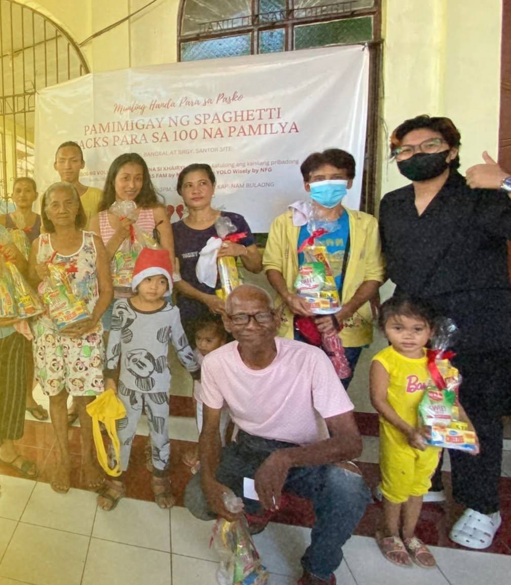 Families and organisers gather under a white banner that reads “Pamimigay ng spaghetti packs para sa 100 na pamilya”, many holding gift bags.