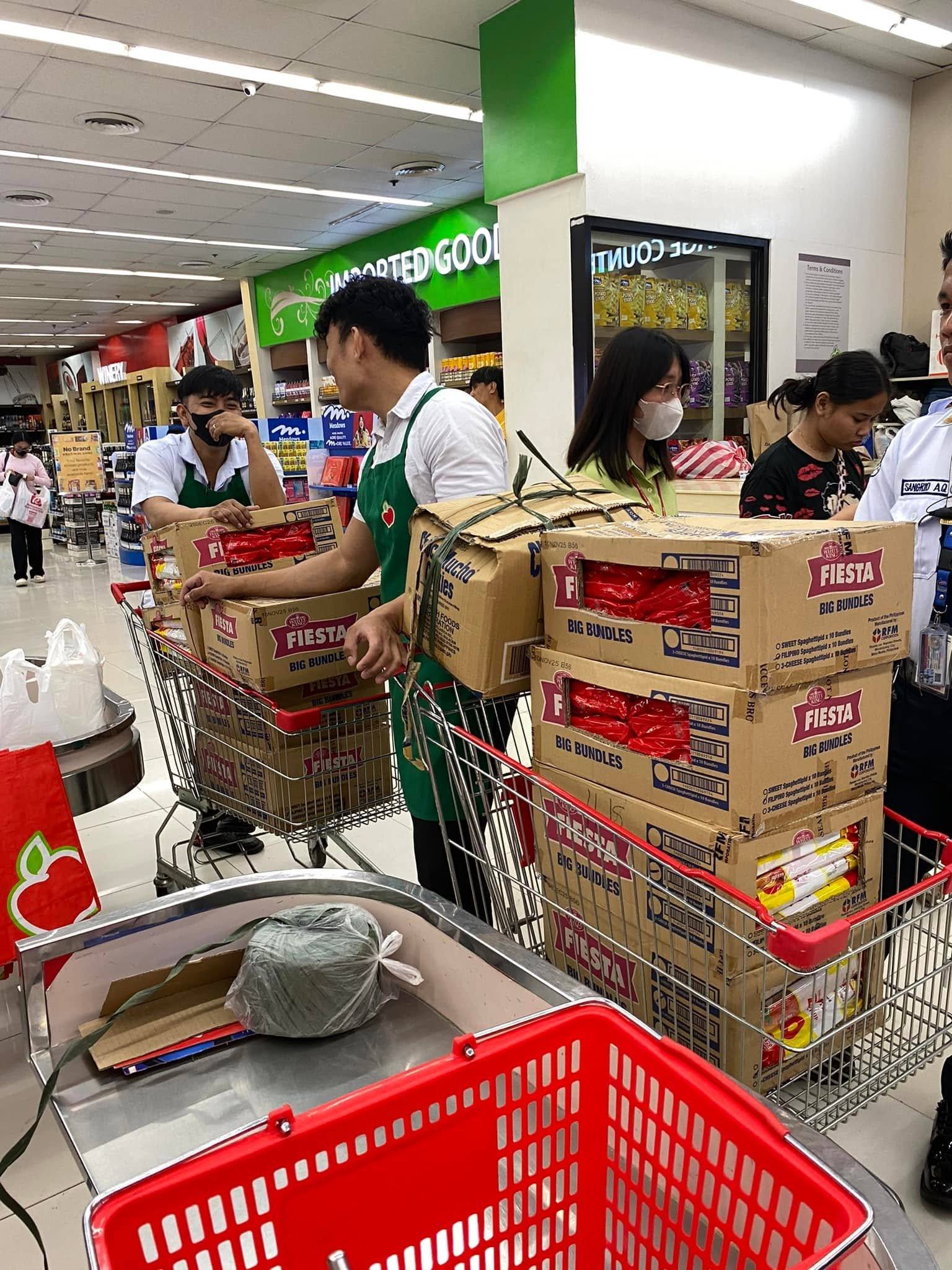 Store staff in green aprons push carts loaded with boxes of Fiesta spaghetti packs toward the checkout.