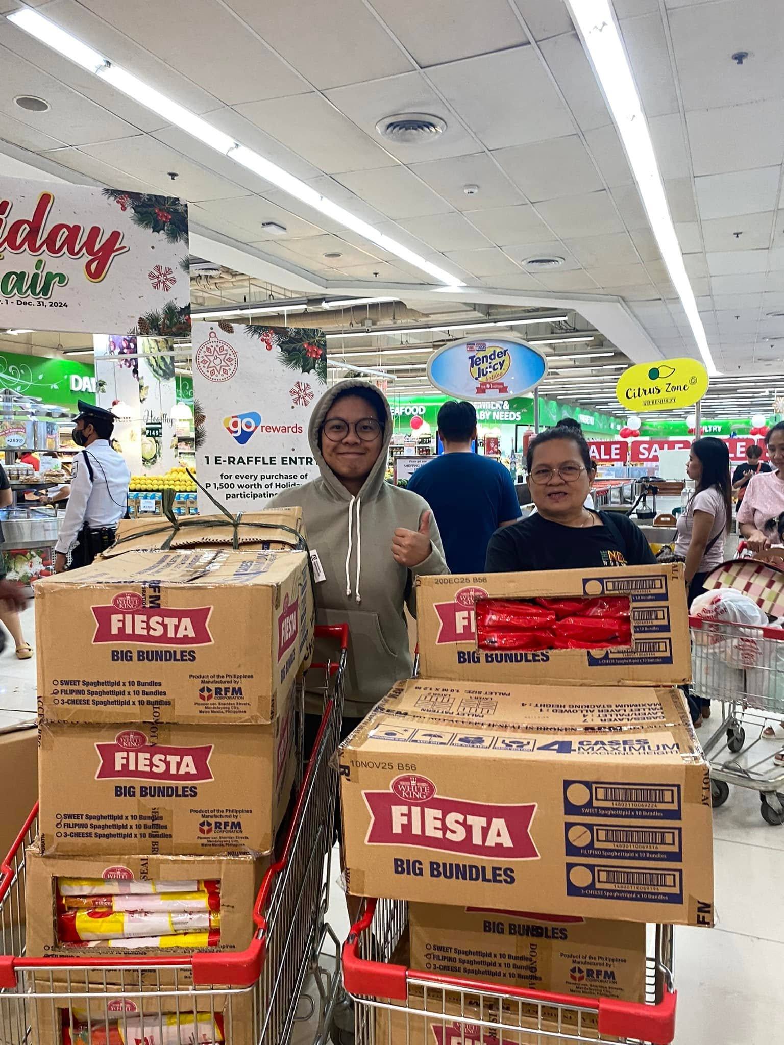 Two people give a thumbs-up beside shopping carts stacked high with boxes of Fiesta spaghetti, in a supermarket decorated for the holidays.
