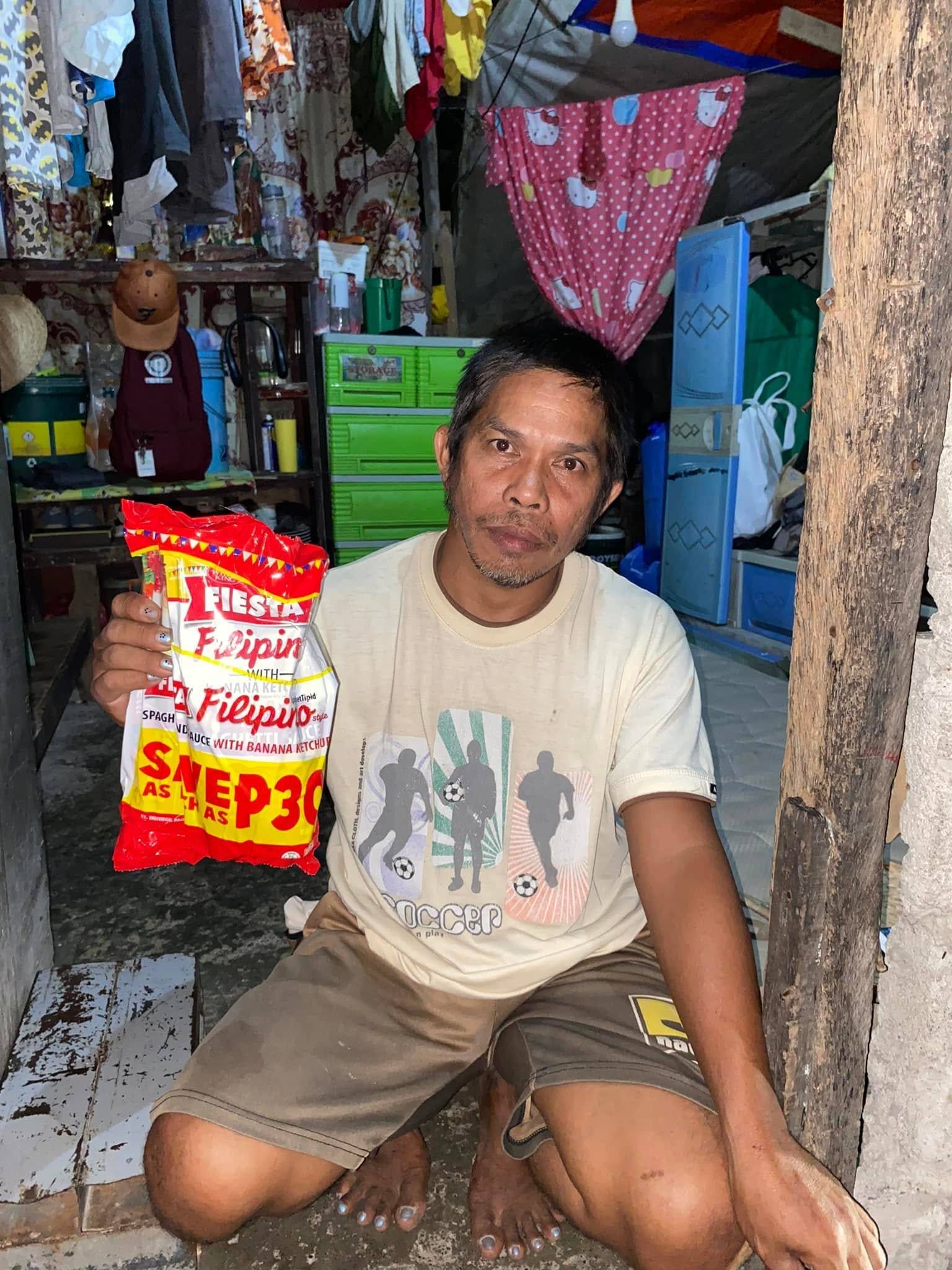 A man crouching in the doorway of his home smiles and holds up a spaghetti pack.