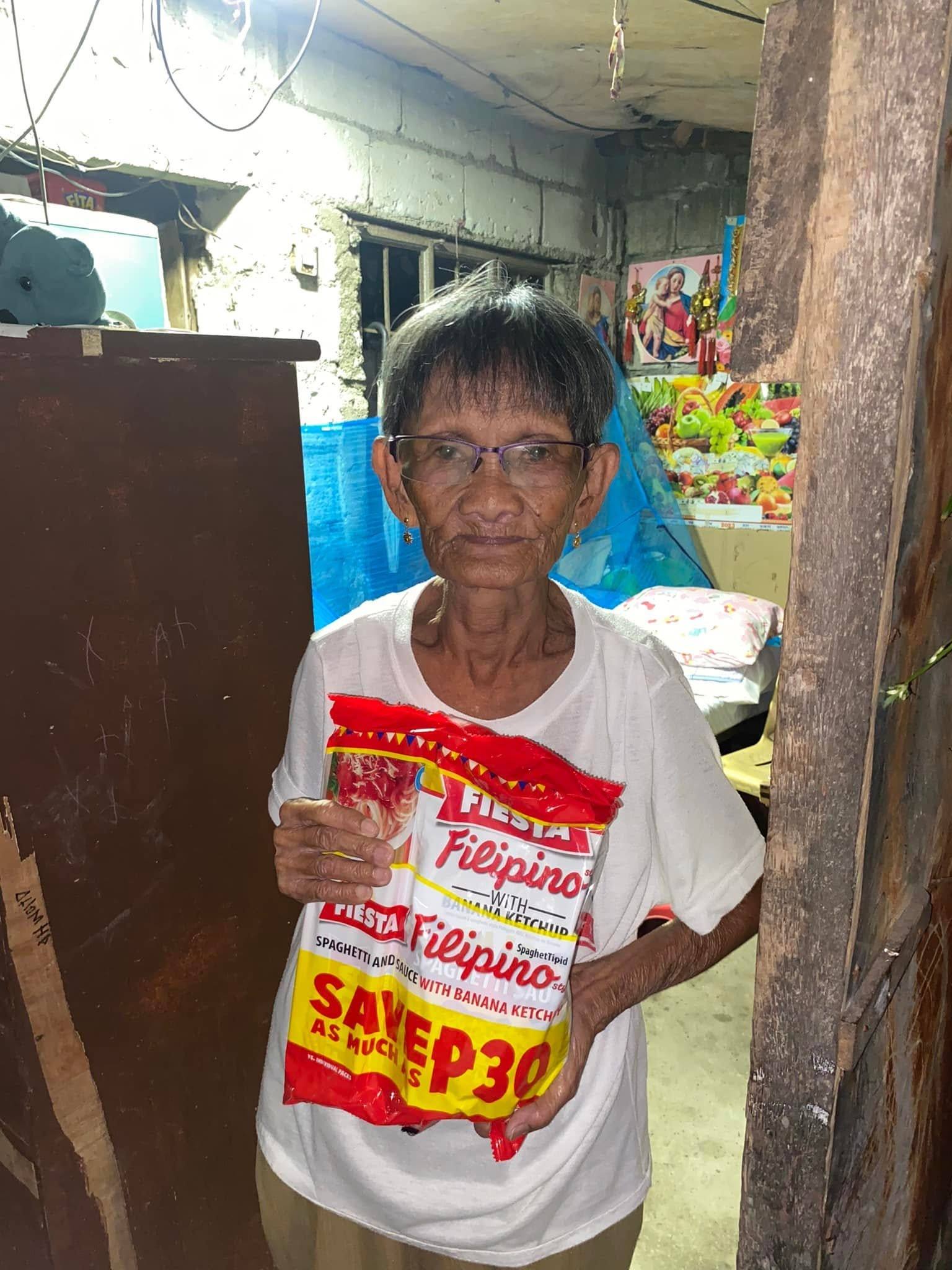 An elderly woman in glasses holds a spaghetti pack at the door of her home.