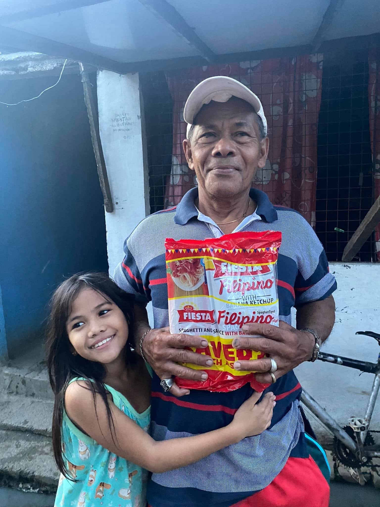 An older man in a cap holds a spaghetti pack while a young girl hugs his arm, both smiling.