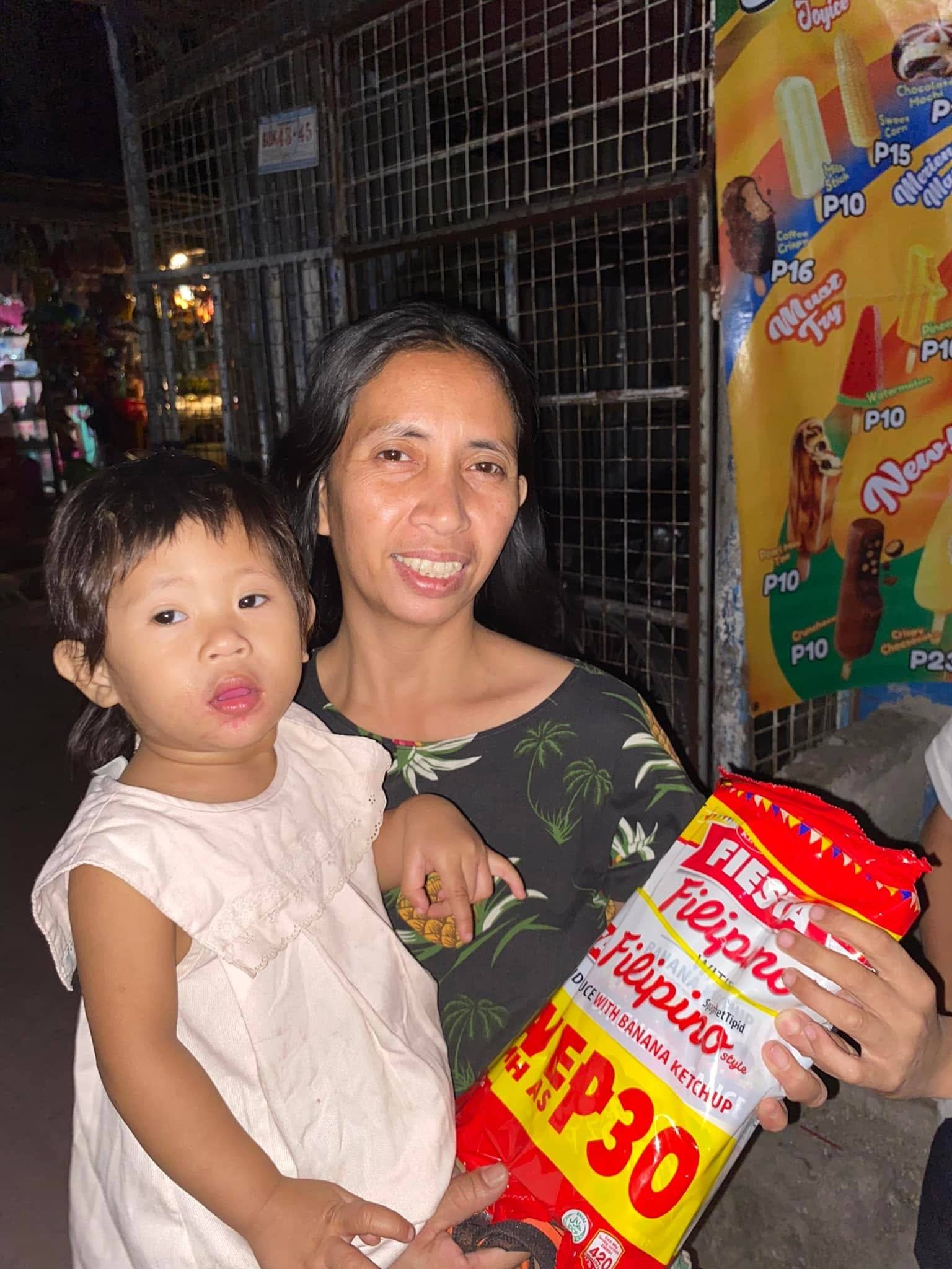 A mother holds her toddler in one arm and a spaghetti pack in the other, at night in front of a small store.