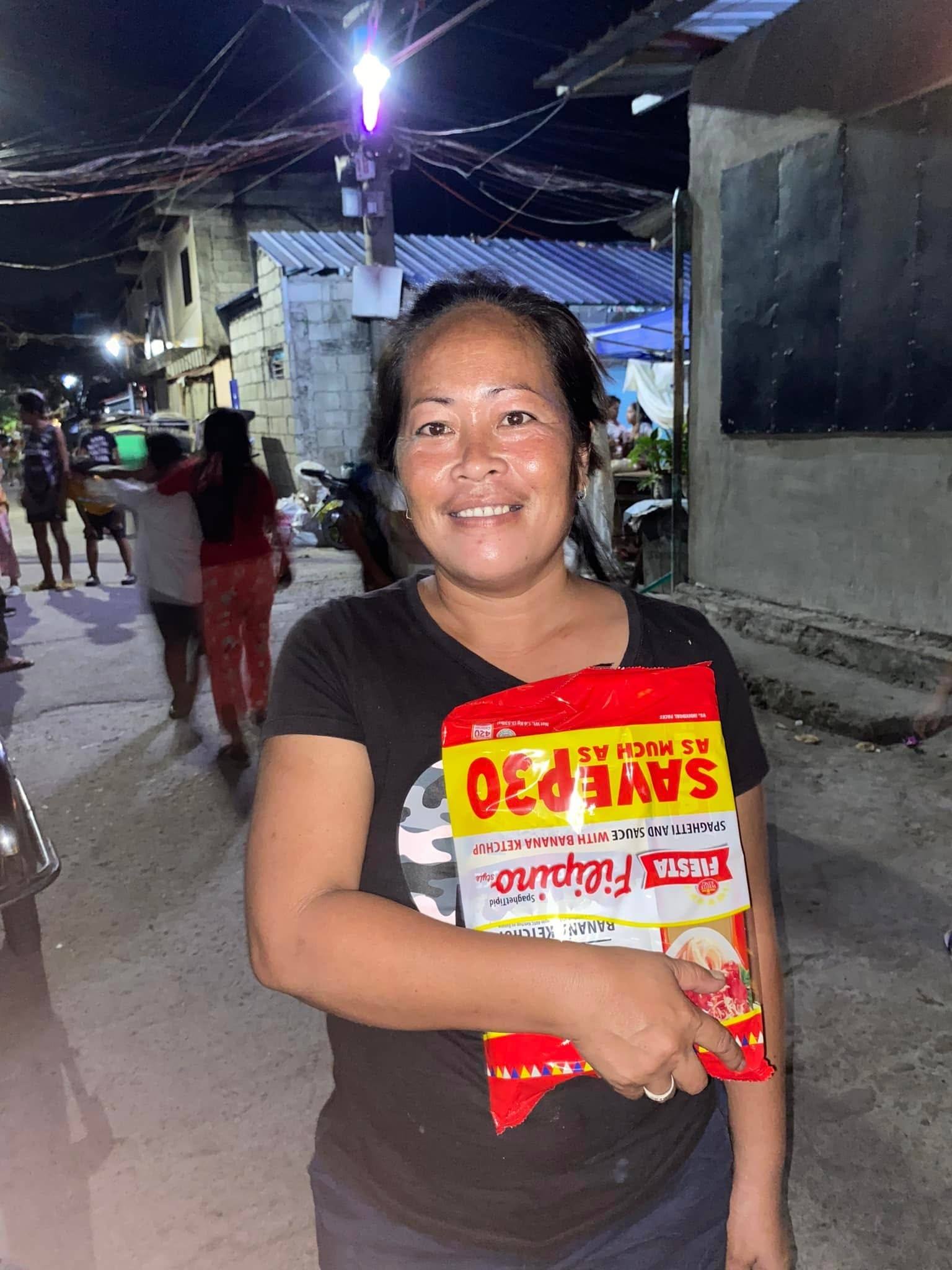 A smiling woman holds her spaghetti pack on a lamplit street at night.