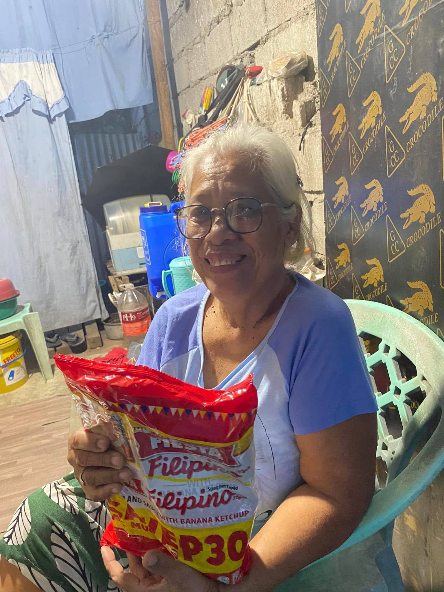 An older woman in glasses, seated in a plastic chair at home, smiles and holds up her spaghetti pack.