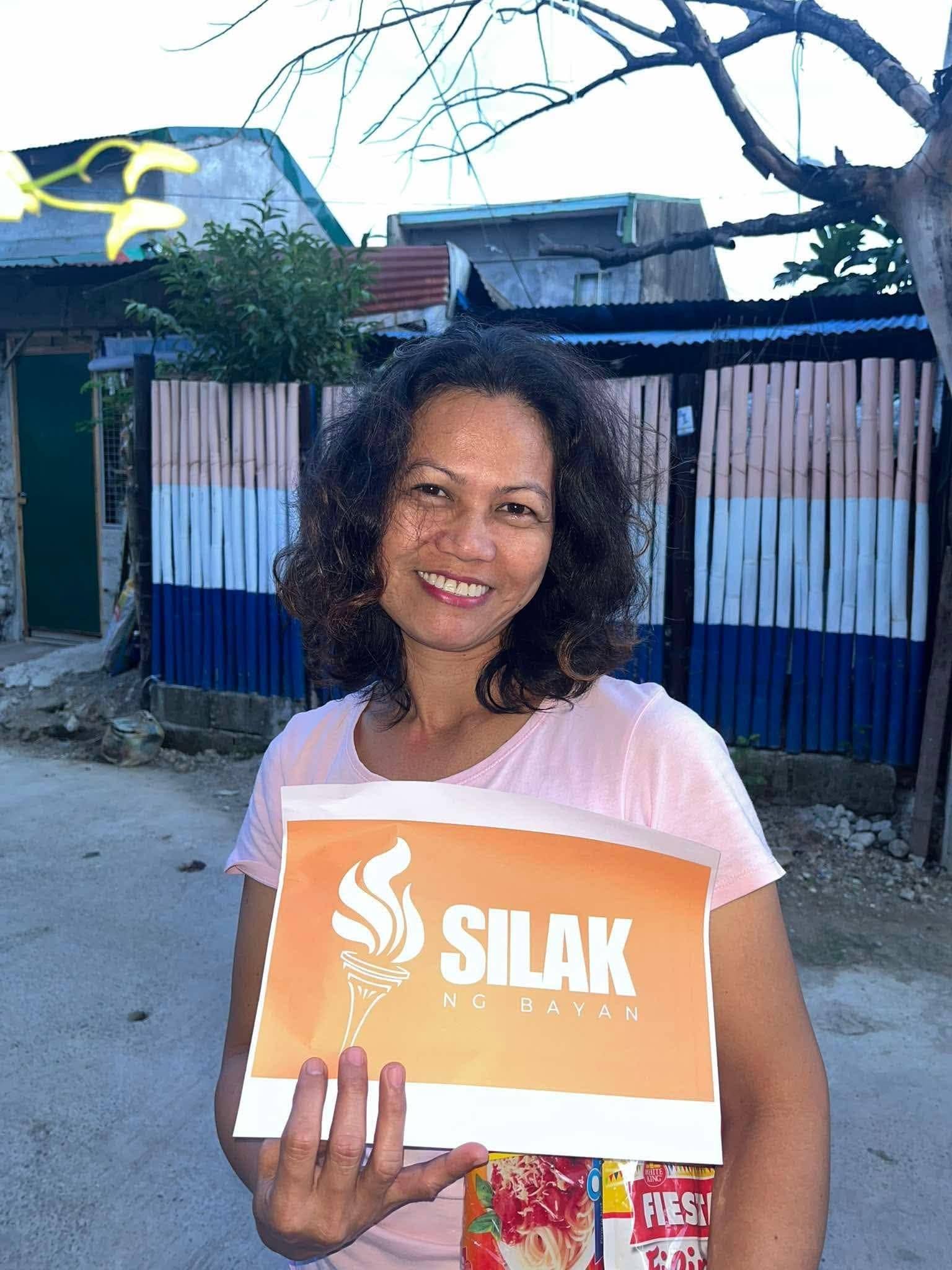 A smiling woman holds up an orange SILAK NG BAYAN sign, her spaghetti pack tucked behind it.