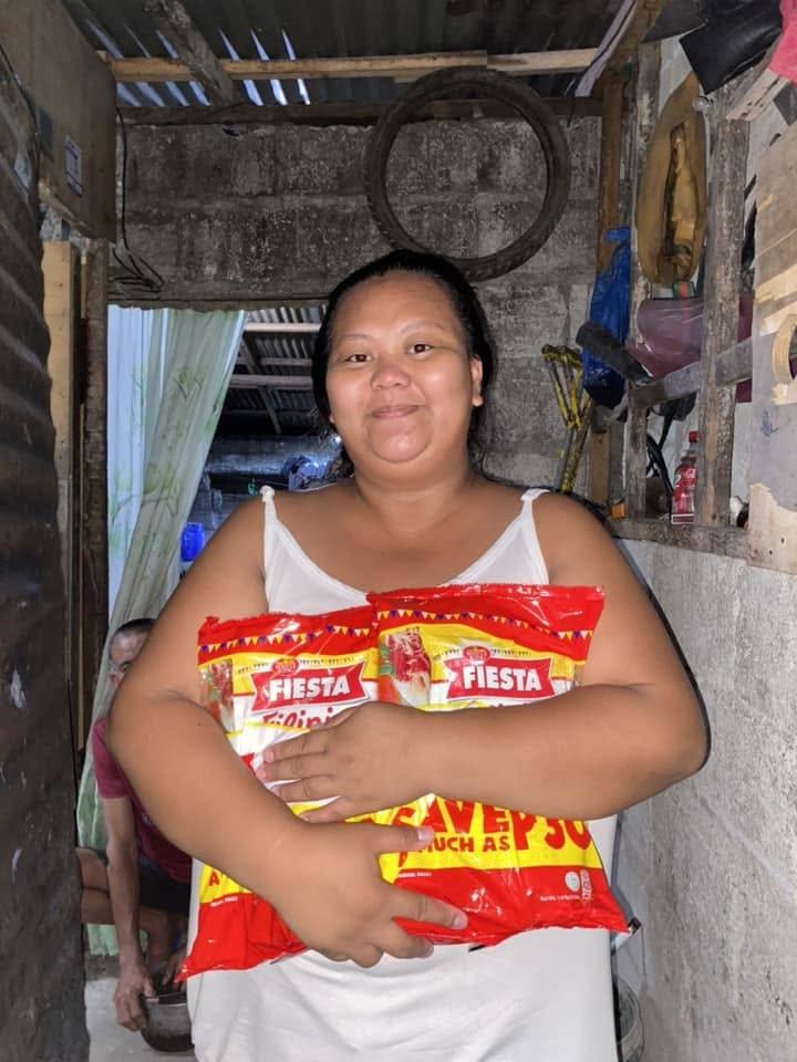 A smiling woman hugs two spaghetti packs to her chest in the doorway of her home.
