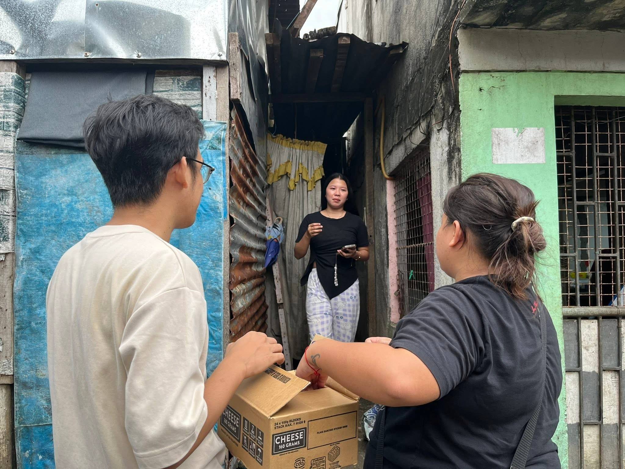 Two people hand out packs from a carton marked “cheese” to a woman at the doorway of her home in a narrow alley.
