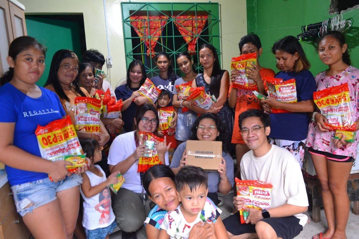 A large group of adults and children pose together with their spaghetti packs in front of a window hung with red Christmas bunting.