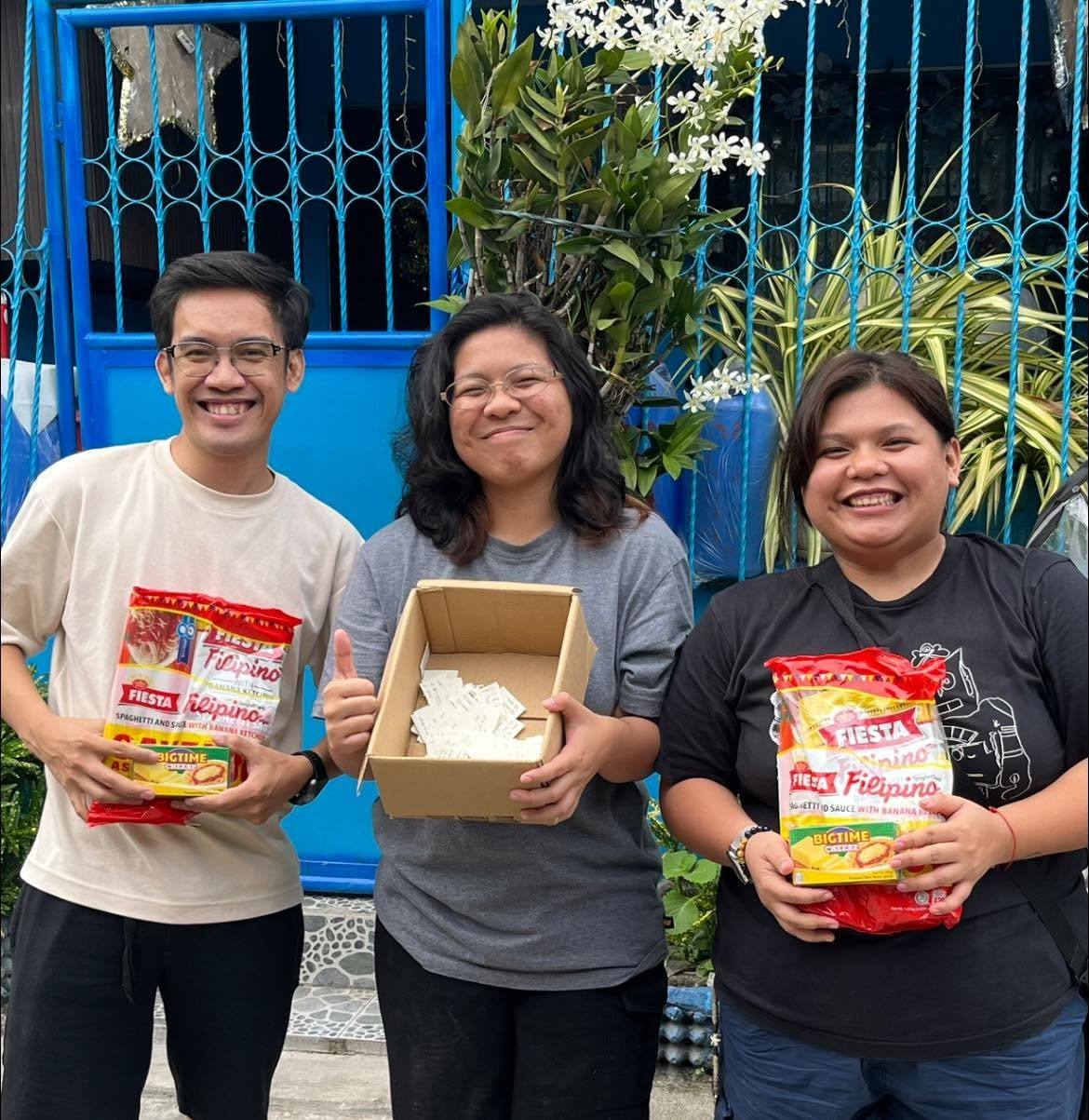 Three smiling people hold spaghetti packs and an open box outside a blue gate.