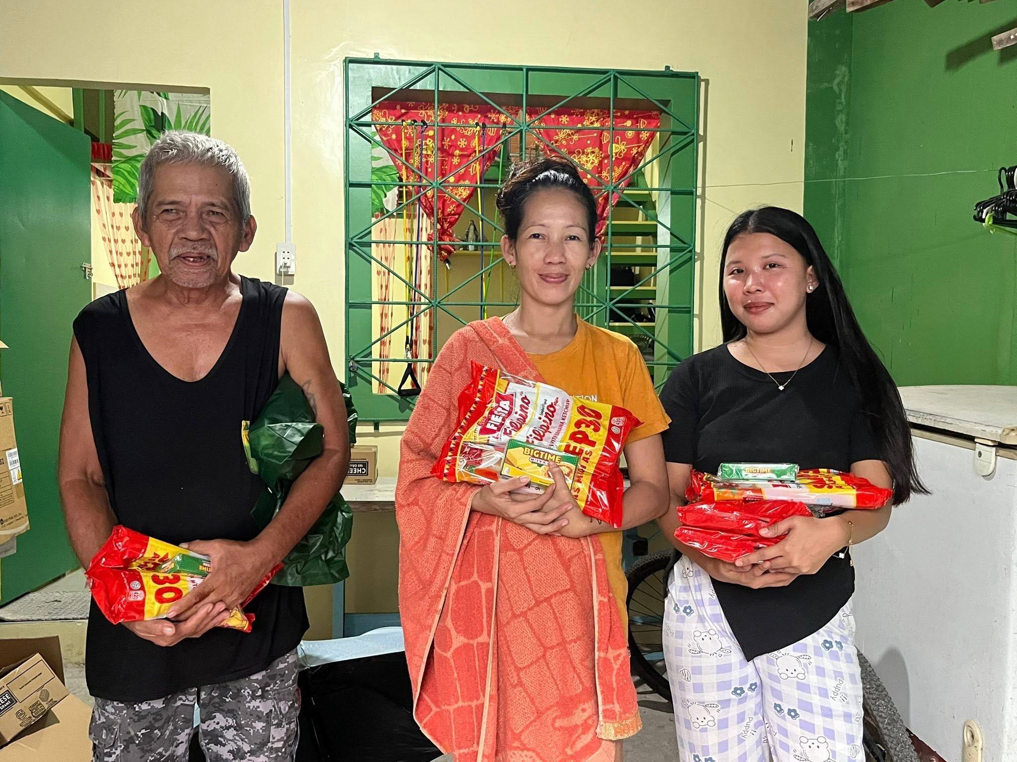 An older man, a woman in an orange top and a young woman stand together holding red spaghetti packs, in a green room with red Christmas bunting on the window grille.