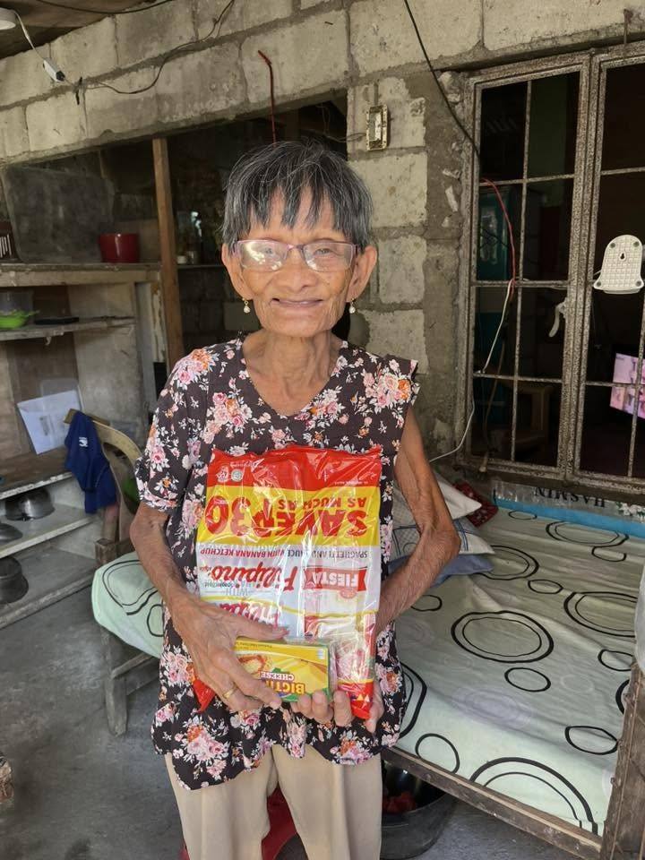 An elderly woman in glasses and a floral blouse smiles inside her home, holding a spaghetti pack.