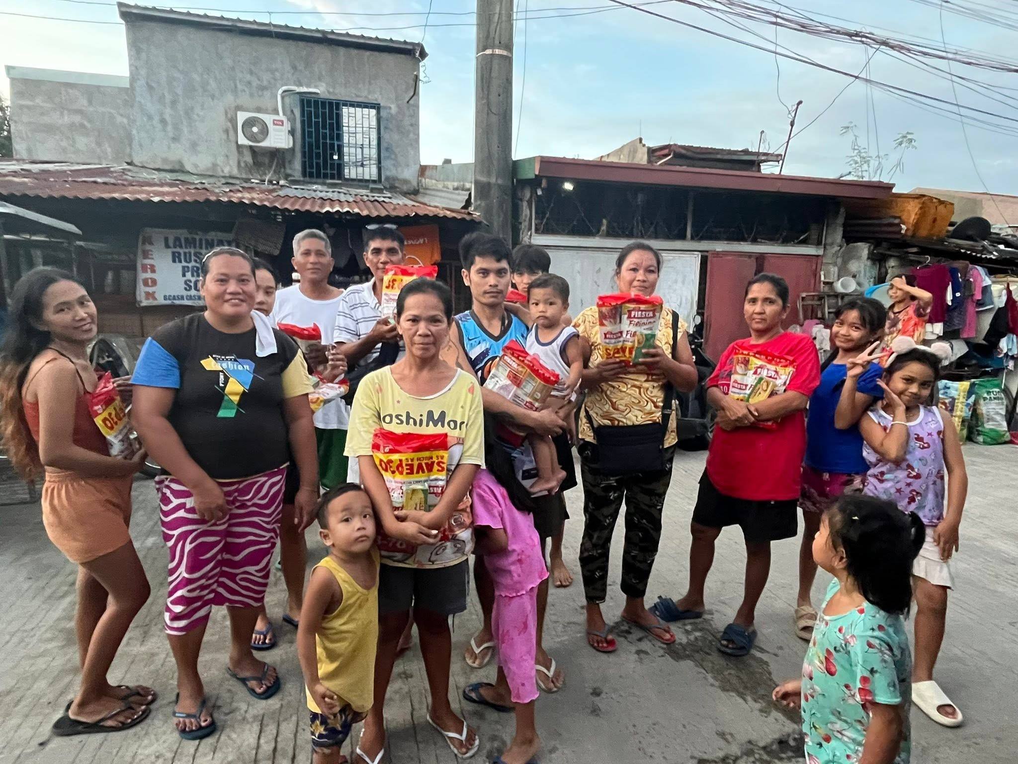 Mothers, fathers and children stand in the street holding their spaghetti packs, a row of houses behind them.
