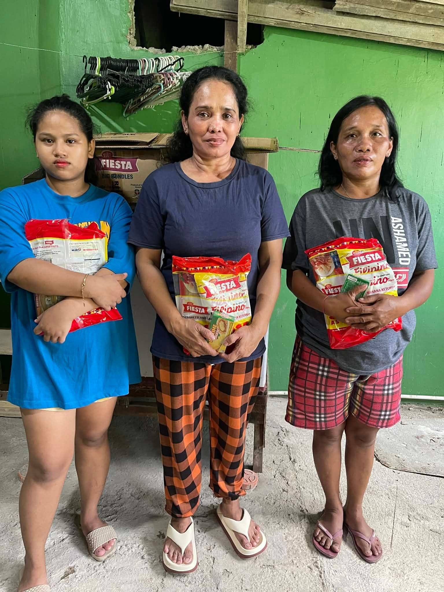 Three women stand side by side against a green wall, each holding a spaghetti pack.