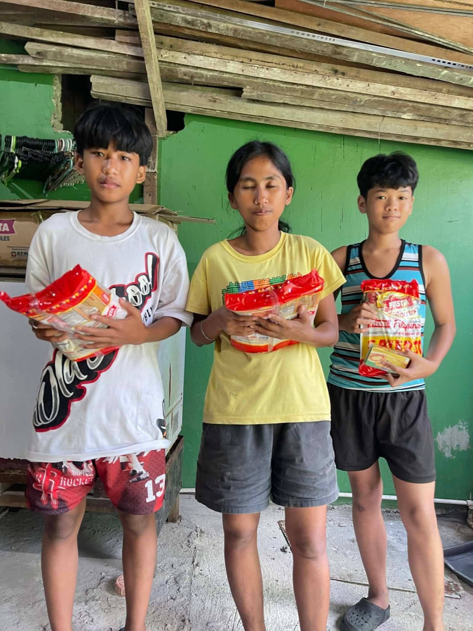 Three young people stand against a green wall, each holding a spaghetti pack.