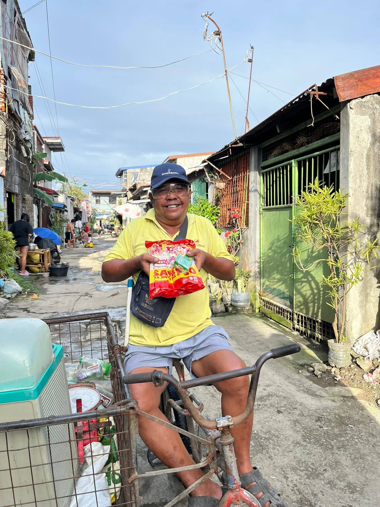 A smiling man in a yellow shirt sits on a bicycle with a sidecar in a narrow street, holding up his spaghetti pack.