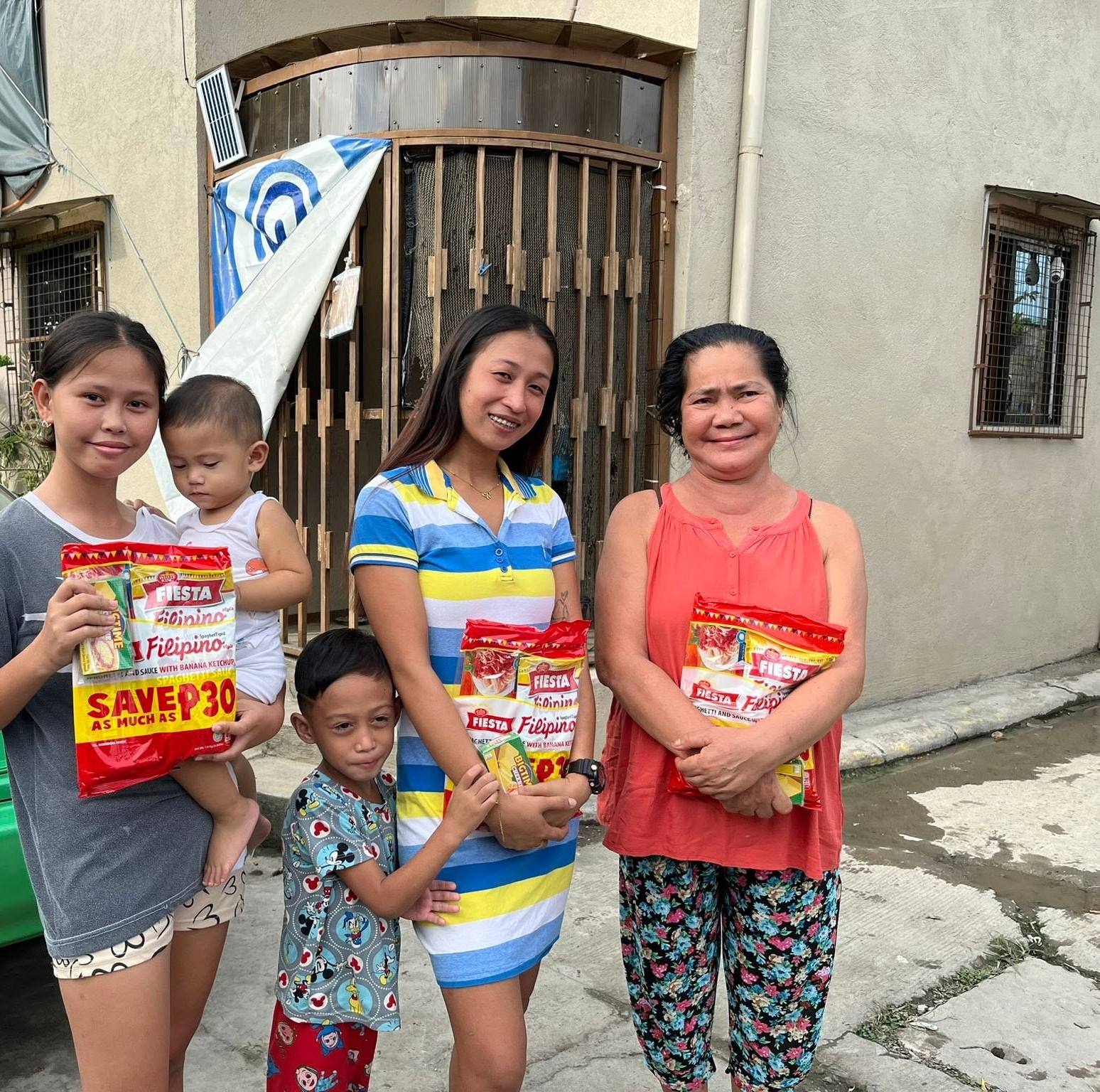 Three women and two small children stand outside a house, holding their spaghetti packs.