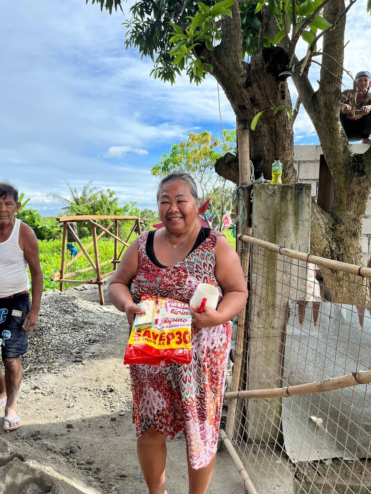 A smiling woman in a patterned dress holds her spaghetti pack by a bamboo fence, green fields and blue sky behind her.