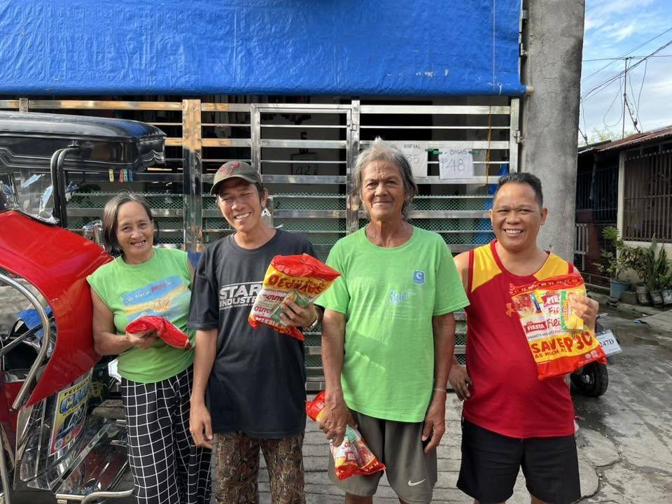 Four neighbours laugh together beside a parked tricycle, packs in hand.