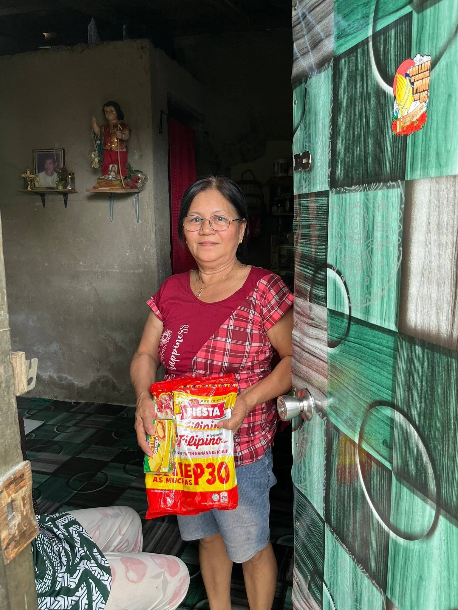 A woman in glasses stands at her open front door holding a spaghetti pack, a small Santo Niño statue on the shelf behind her.