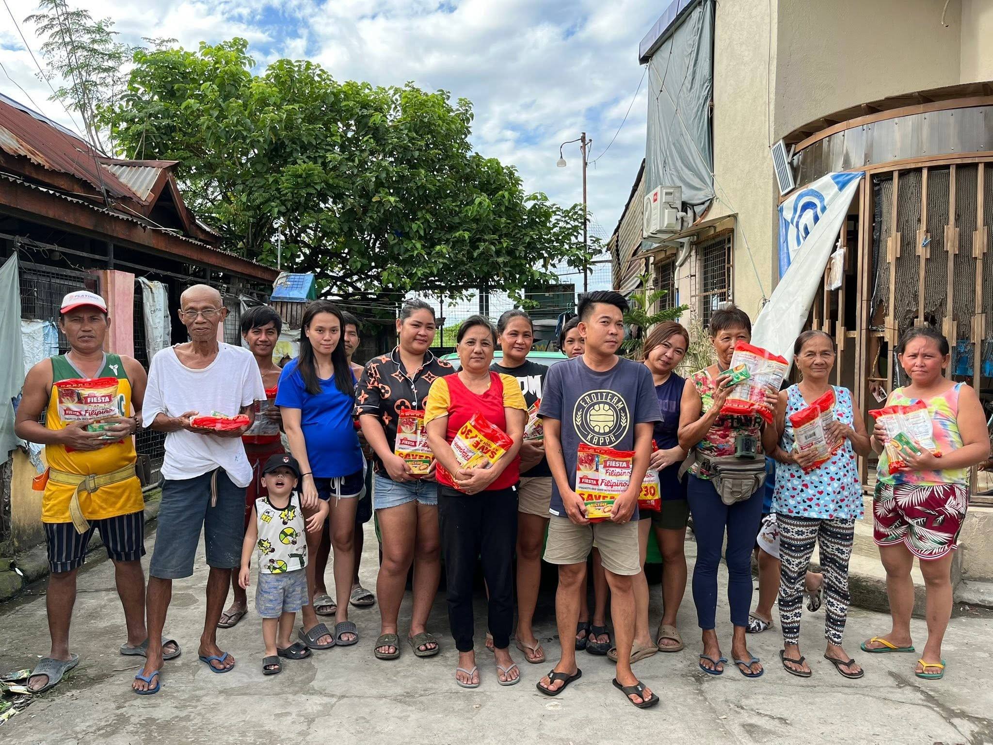 About twenty residents, young and old, stand in a line on a village street under a bright sky, holding their spaghetti packs.